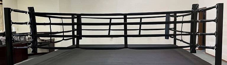 Elevated commercial boxing ring with black canvas, ropes, corner cushions, and skirt